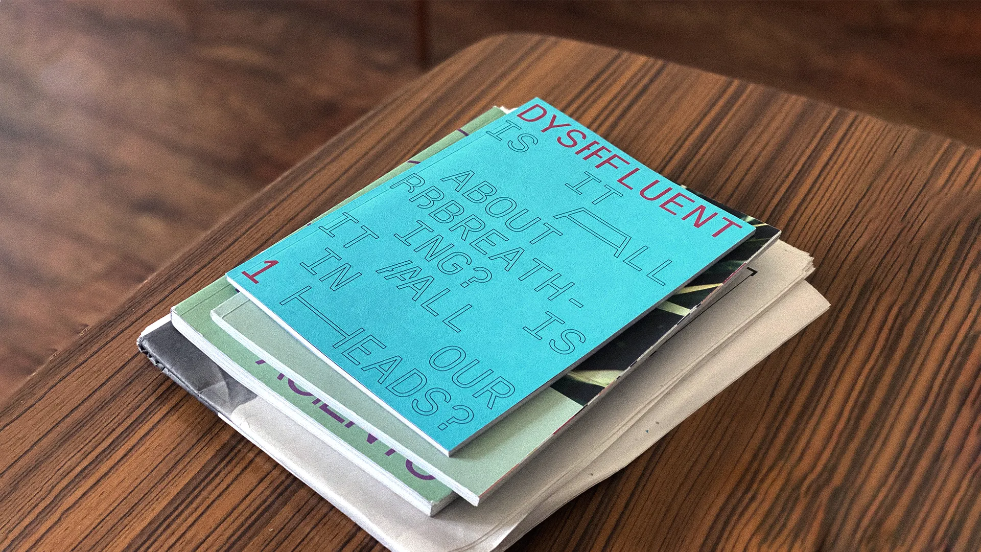 A stack of magazines on a wooden table. Dysfluent magazine issue 1 is at the top. It is cyan-coloured and its title ‘Dysfluent 1’ in bright red. 