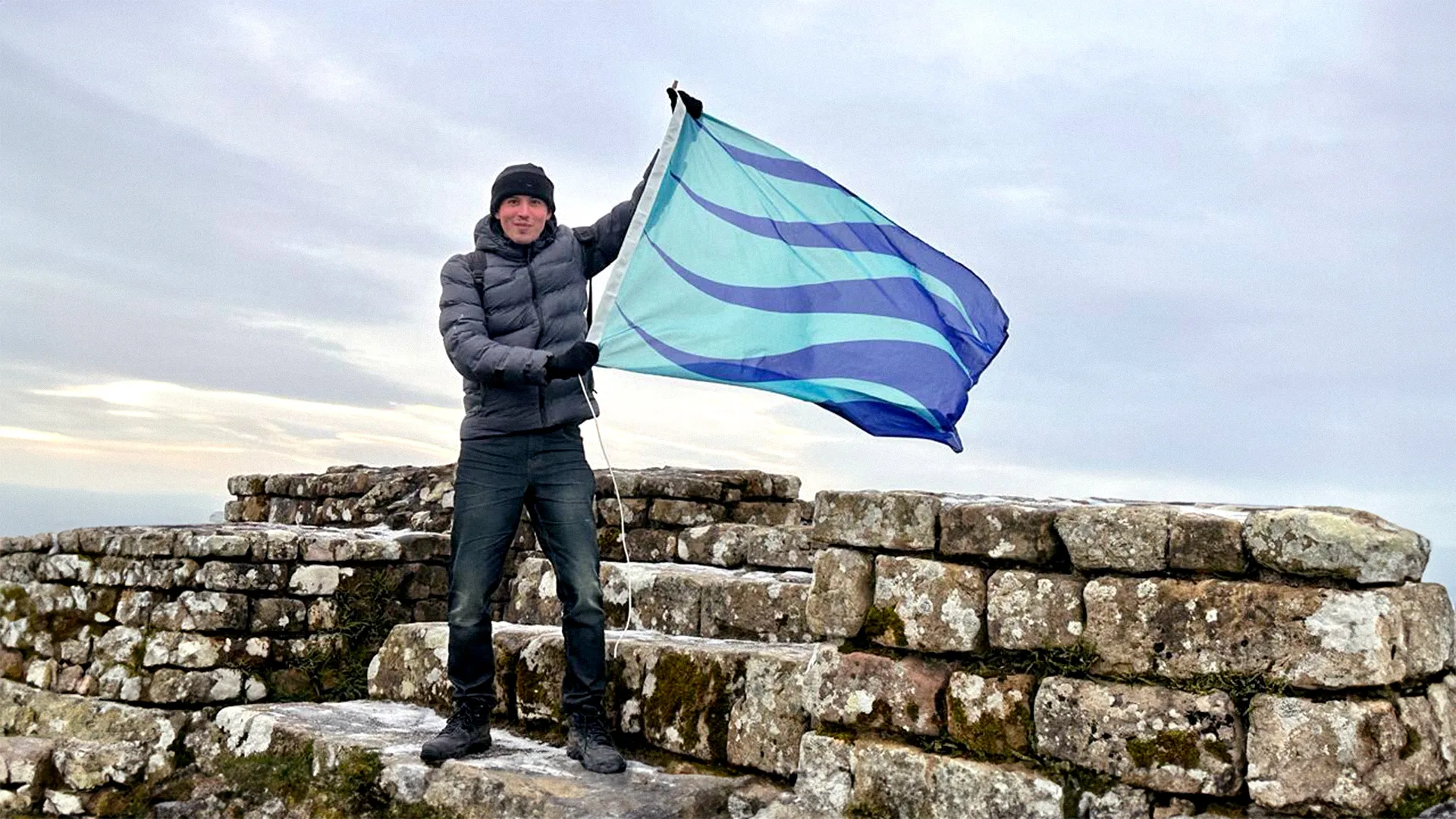 A man wearing a winter coat standing on a ruined wall holding a flag with his two hands. The flag is seagreen and ultramarine blue waves.
