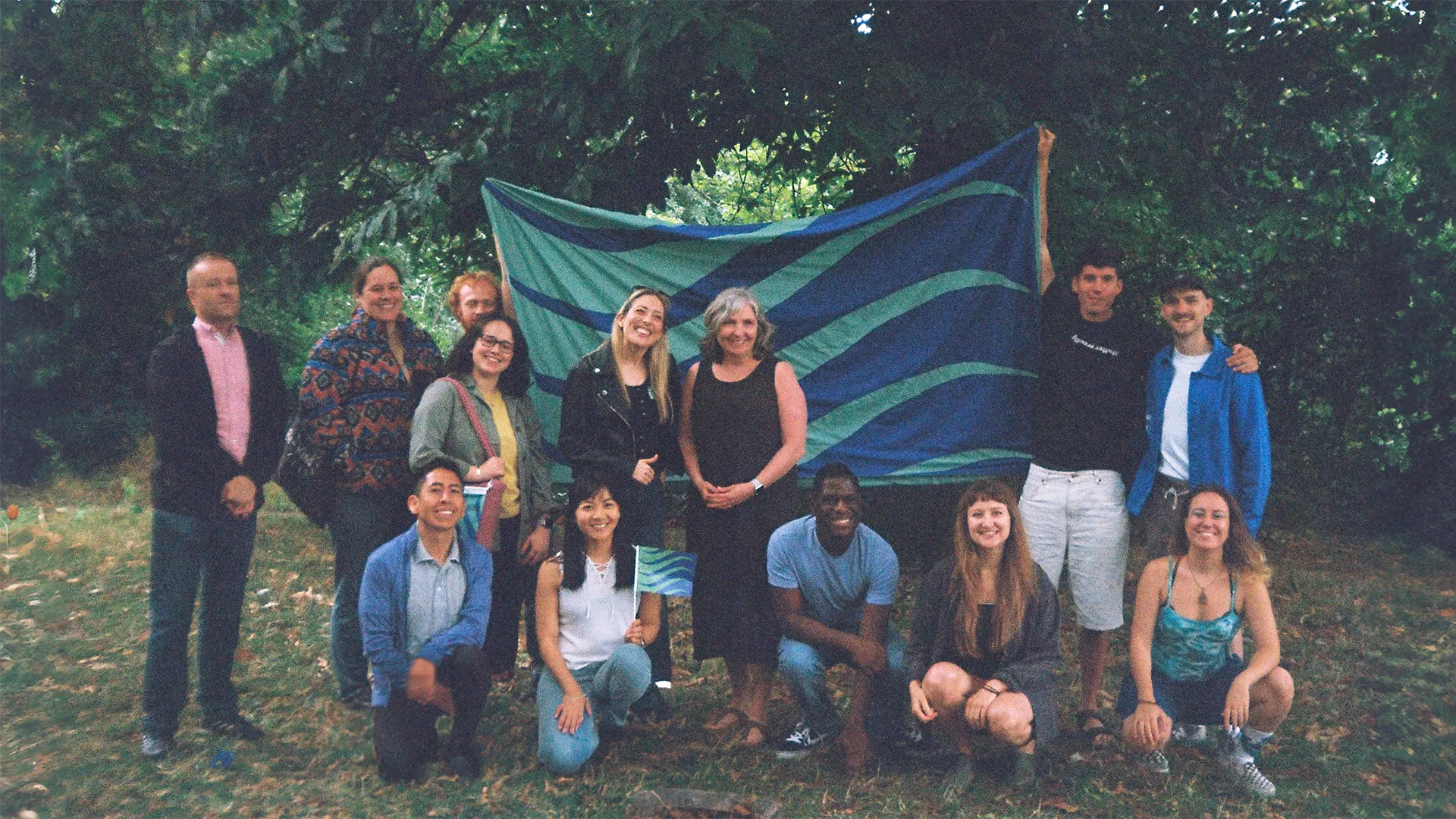 A group of people standing in a wooded, grassy area. The group is diverse in age, skin colour, body size and clothing colour and texture. In the centre of the composition, two people are holding a large, hand-made flag. The flag is a stuttering pride flag, and features seagreen and ultramarine waves.