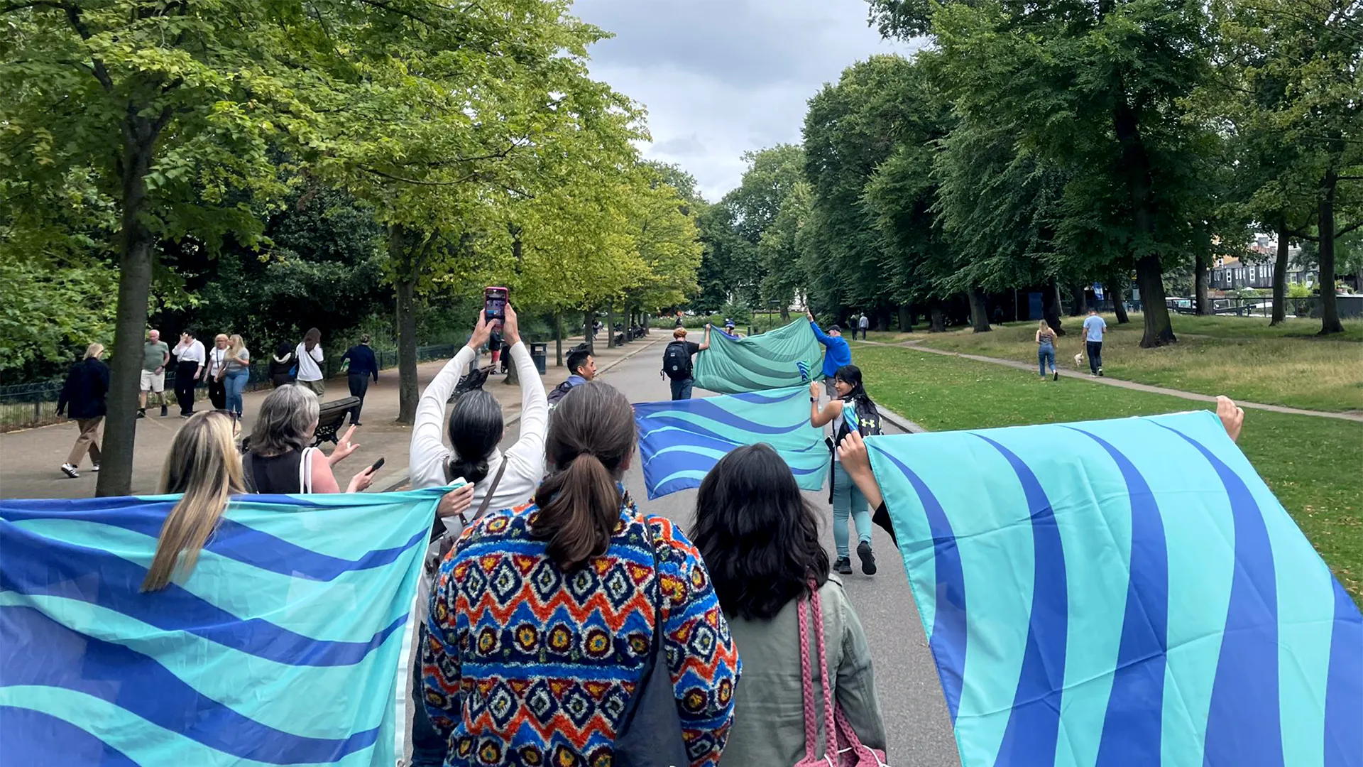A group of people march down a wide park pathway, waving flags. The flags are seagreen and ultramarine blue waves.
