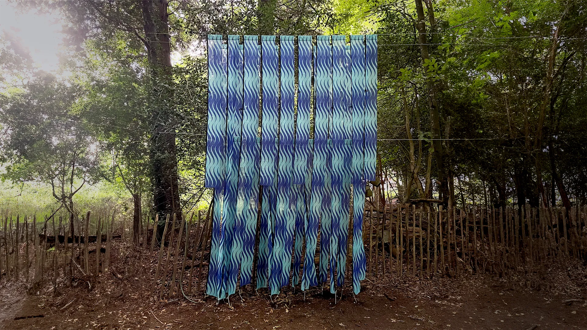 A sculpture in a forest. It is made of long strips of a repeat pattern on flag material. The repeat design is seagreen and ultramarine blue waves. 