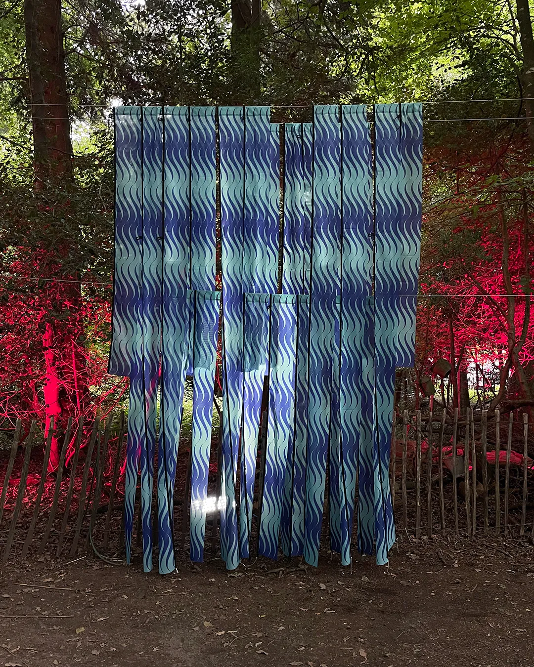 A sculpture in a forest. It is made of long strips of a repeat pattern on flag material. The repeat design is seagreen and ultramarine blue waves. It is dark and the sculpture is backlit with lights.