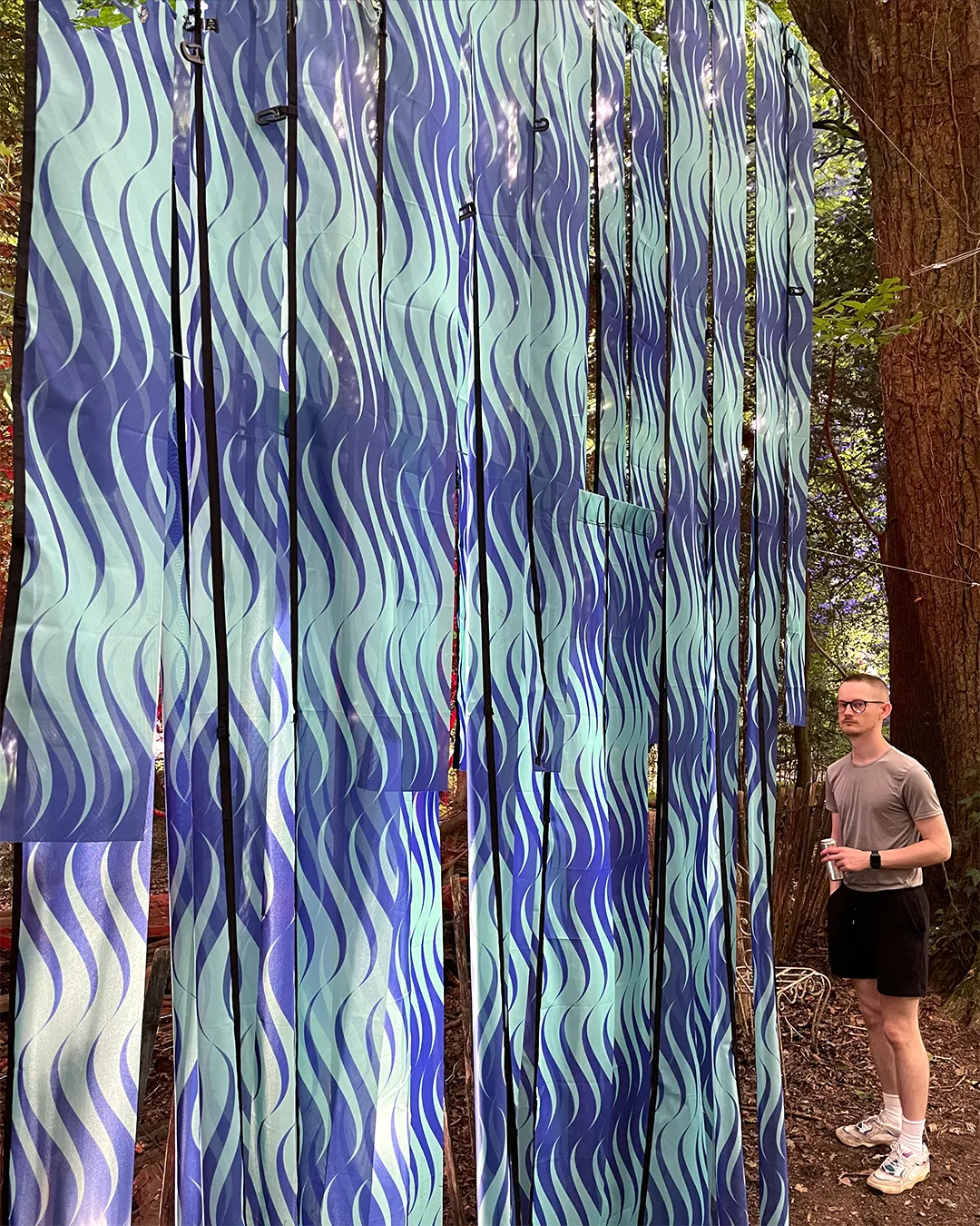 A sculpture in a forest. It is made of long strips of a repeat pattern on flag material. The repeat design is seagreen and ultramarine blue waves. A man stands to the side.