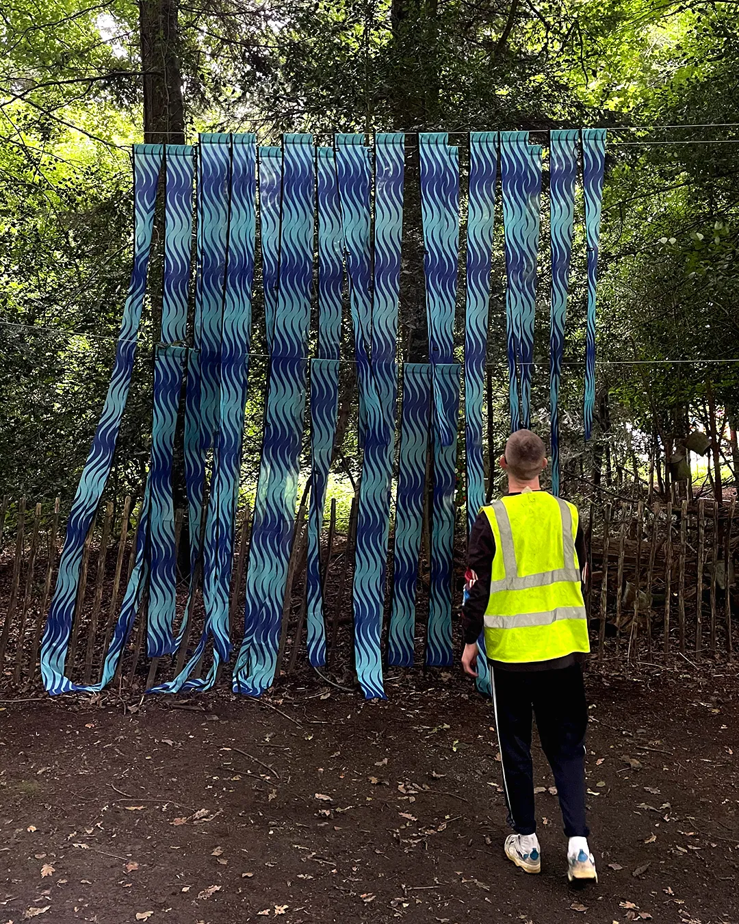 A sculpture in a forest. It is made of long strips of a repeat pattern on flag material. The repeat design is seagreen and ultramarine blue waves. A man in a  high-vis jacket stands in front.