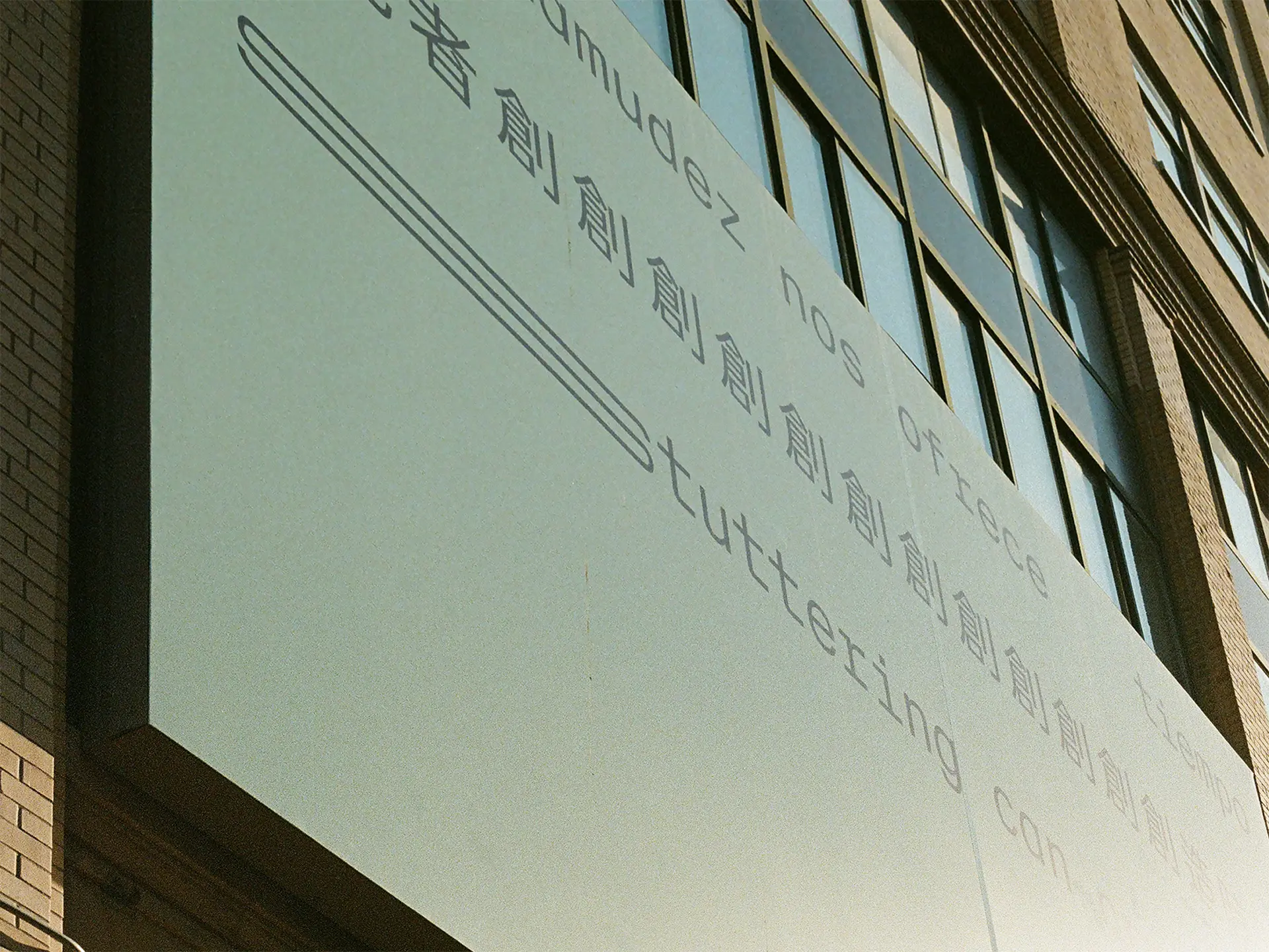 A close-up photograph of sun shining on a billboard.  Three lines of black text appear on a light seafoam green background, in Spanish, Chinese and English, translating to: ‘Stuttering can create time.’ The text is in a sans serif typeface organised in three straight lines within the top half of the composition. The bottom half of the composition is empty. The text is stretched and repeated to represent stammering.