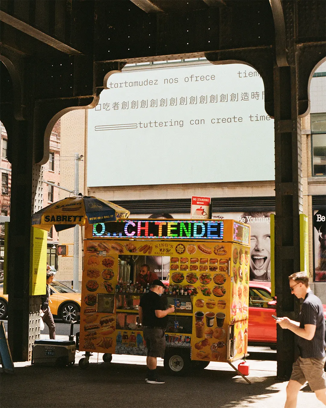 A film photograph of the billboard, as seen from ground level underneath the High Line. A food truck is in the foreground. 