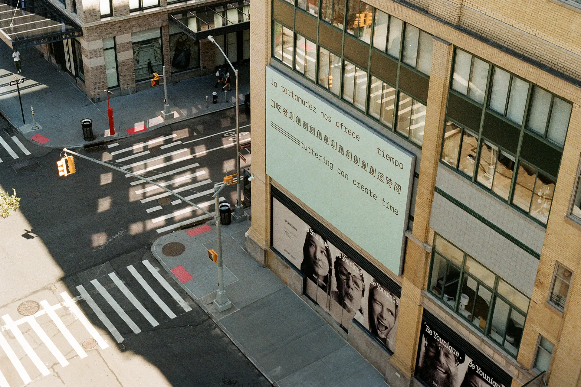 A billboard on a New York City building. Three lines of black text appear on a light seafoam green background, in Spanish, Chinese and English, translating to: ‘Stuttering can create time.’ The text is in a sans serif typeface organised in three straight lines within the top half of the composition. The bottom half of the composition is empty. The text is stretched and repeated to represent stammering. A young man on ground level is looking up at the billboard.