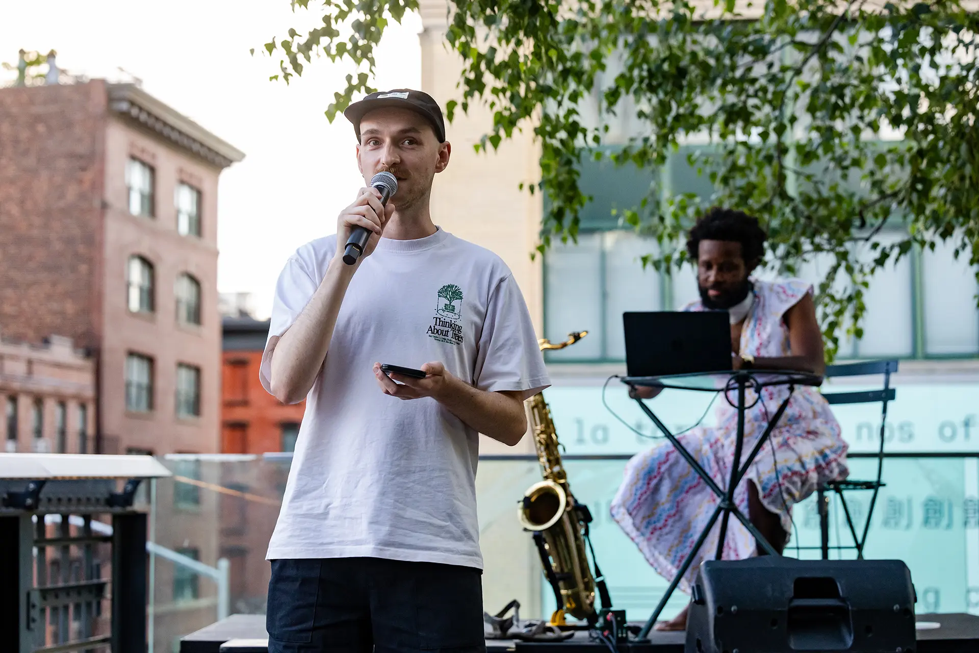 A man wearing a white t-shirt and a black cap speaks into a microphone. 