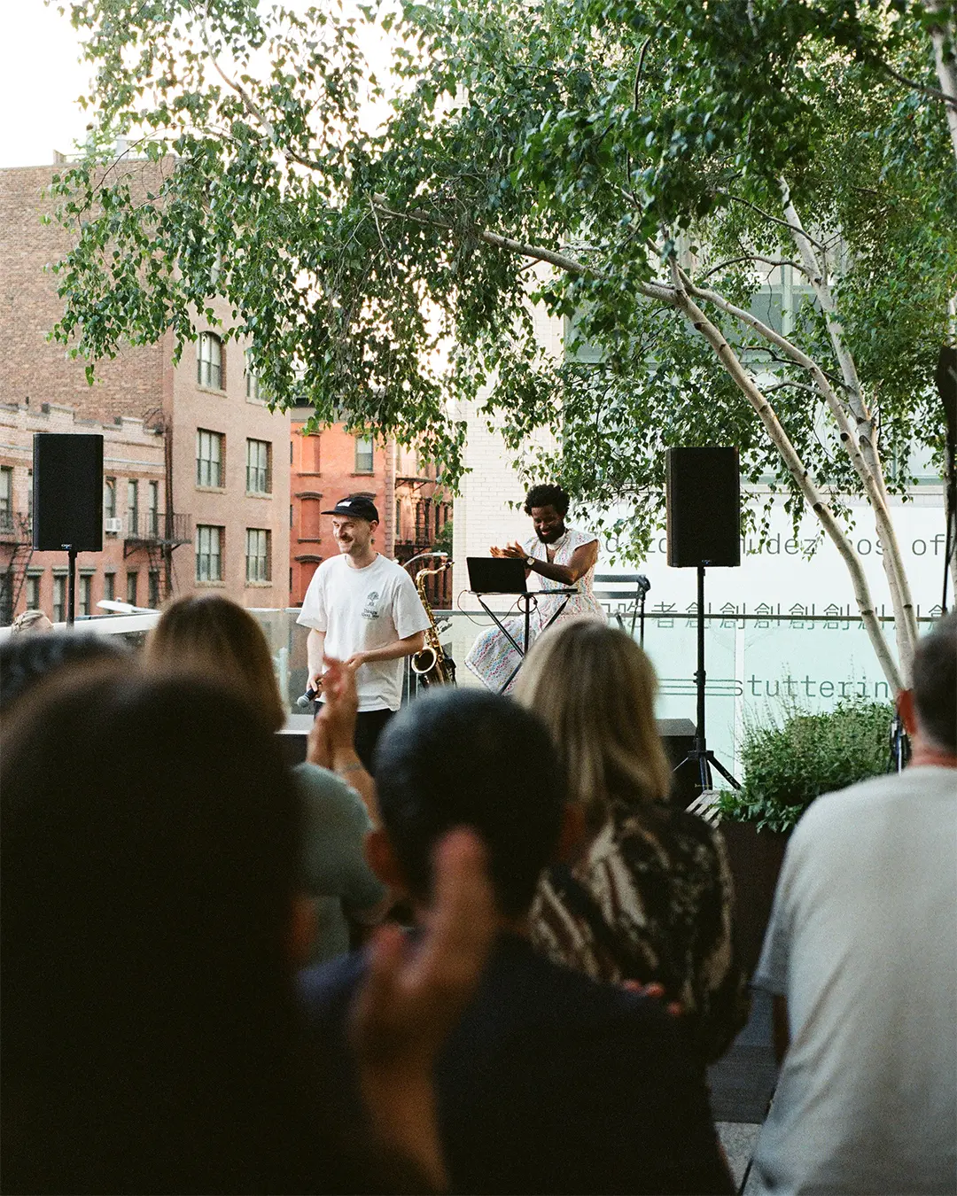 A man wearing a white t-shirt and a black cap speaks into a microphone. The crowd is clapping.