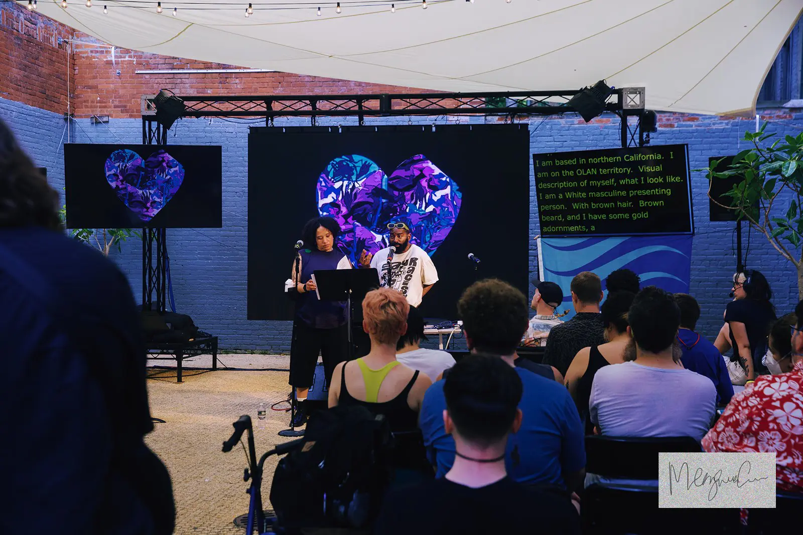 A crowd looks forward at two people presenting. Multiple screens show IWBWYE’s logo, a purple-blue heart. 