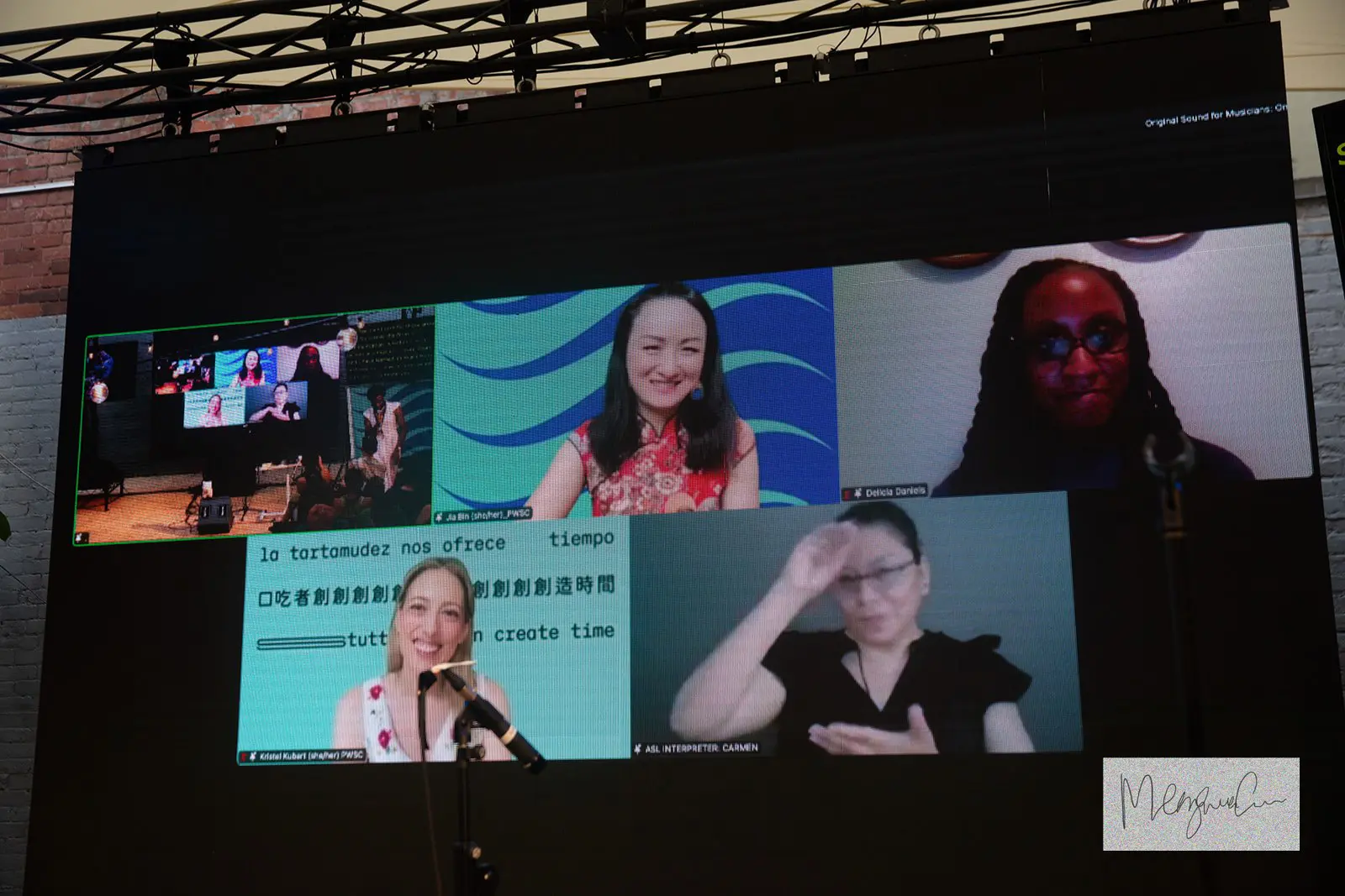 
A photo of a massive screen on stage, showing five video call boxes with people smiling. 
