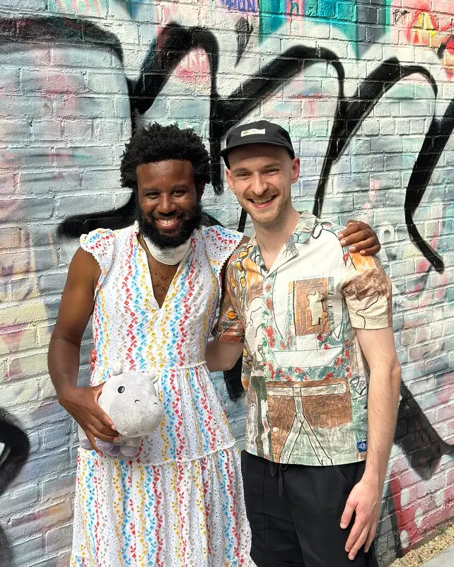 Two people stand and smile in embrace. The person on the left has brown skin and is wearing a colourful dress. The person on the right is light skinned and is wearing a colourful shirt and a black cap. 