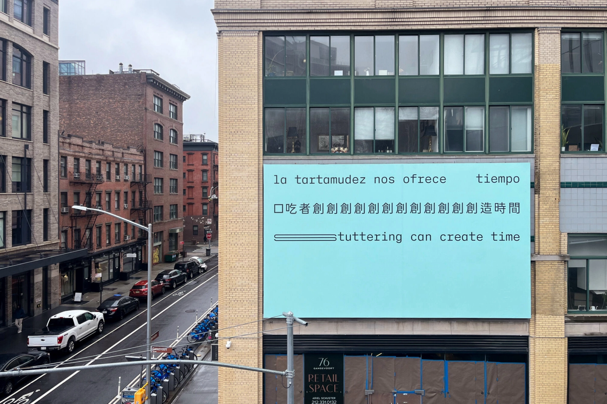 A billboard on a New York City building. Three lines of black text appear on a light seafoam green background, in Spanish, Chinese and English, translating to: ‘Stuttering can create time.’ The text is in a sans serif typeface organized in three straight lines within the top half of the composition. The bottom half of the composition is empty. The text is stretched and repeated to represent stammering.