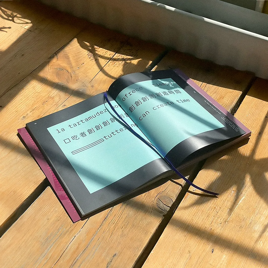A large book on a wooden table, open to show the design of the billboard by People Who Stutter Create. It is green-blue with black typography.