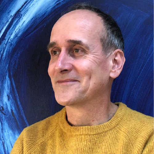 A portrait of Paul Aston: he is wearing a bright yellow jumper. He is looking off into the distance with a smile, his face turned at a three-quarters angle against the camera. Behind him is a painterly blue backdrop.